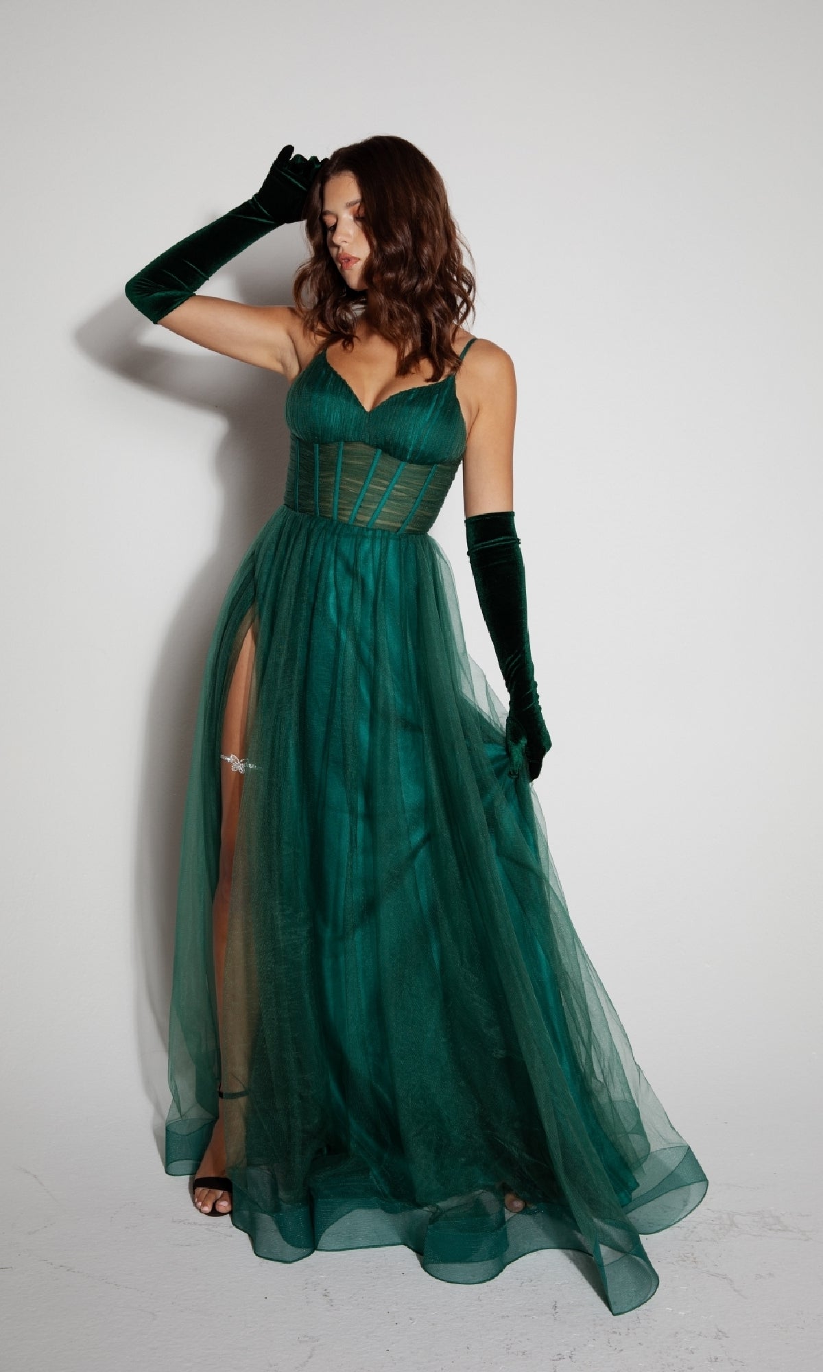 A woman in the Eureka 9177 Corset Long Tulle Prom Ball Gown, an elegant green a-line dress with a corset bodice and matching velvet gloves, stands against a plain white wall.
